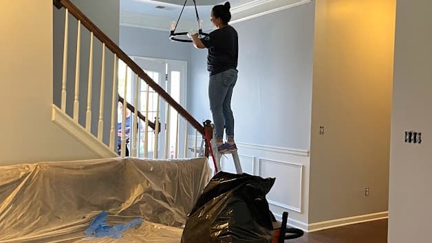 Worker on a ladder painting a two-story entry with the floor and stairs covered in plastic