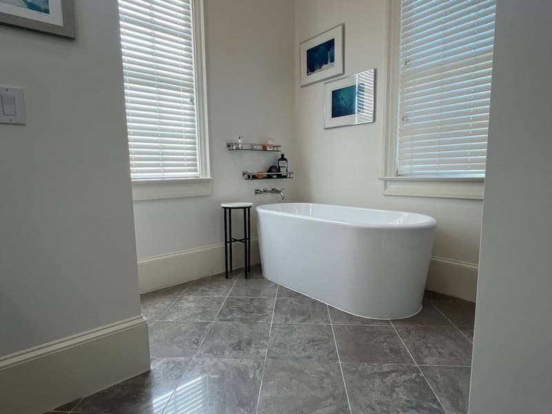 Freestanding white soaking tub below plantation shutters on a grey tile floor